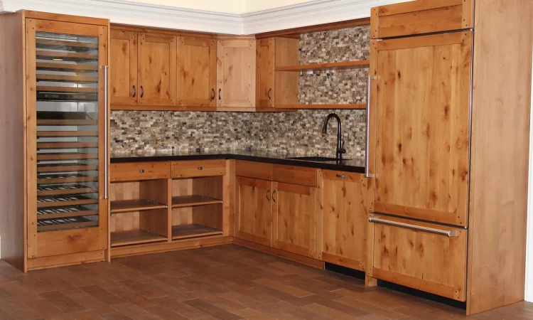 A clean and organized kitchen with ample storage space. Light colored wooden cabinets with brushed nickel handles line the walls. A stainless steel refrigerator stands to the left of the image. A black countertop and sink with faucet are visible in the foreground. Light colored tiles create a backsplash that rises to meet the light colored walls.