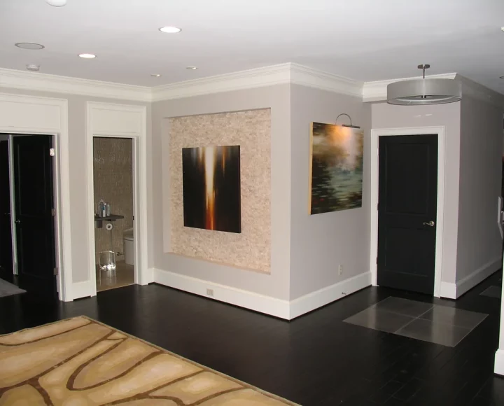 A dimly lit basement hallway with recessed lights on the ceiling. Two paintings hang on the right wall: a vibrant abstract piece and a black and white landscape. A patterned rug warms the cold concrete floor.