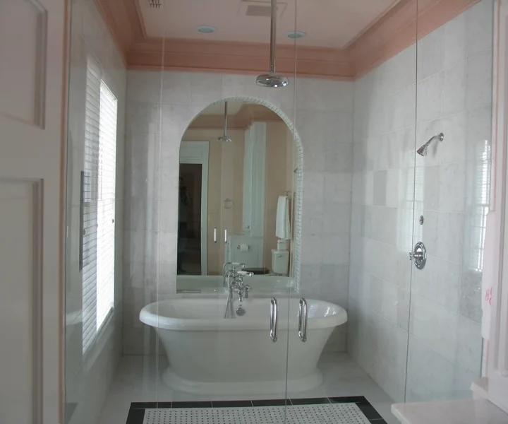 A bathroom with a fixed bathtub enclosed in a glass shower door. The bathroom walls are tiled in a light color, and a shower tile floor with a decorative border. There's a mirror behind the bathtub.