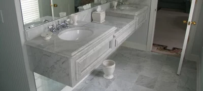 A bright bathroom with two rectangular sinks with chrome faucets mounted on a marble countertop with drawers. A large rectangular mirror is mounted on the wall above the sinks.